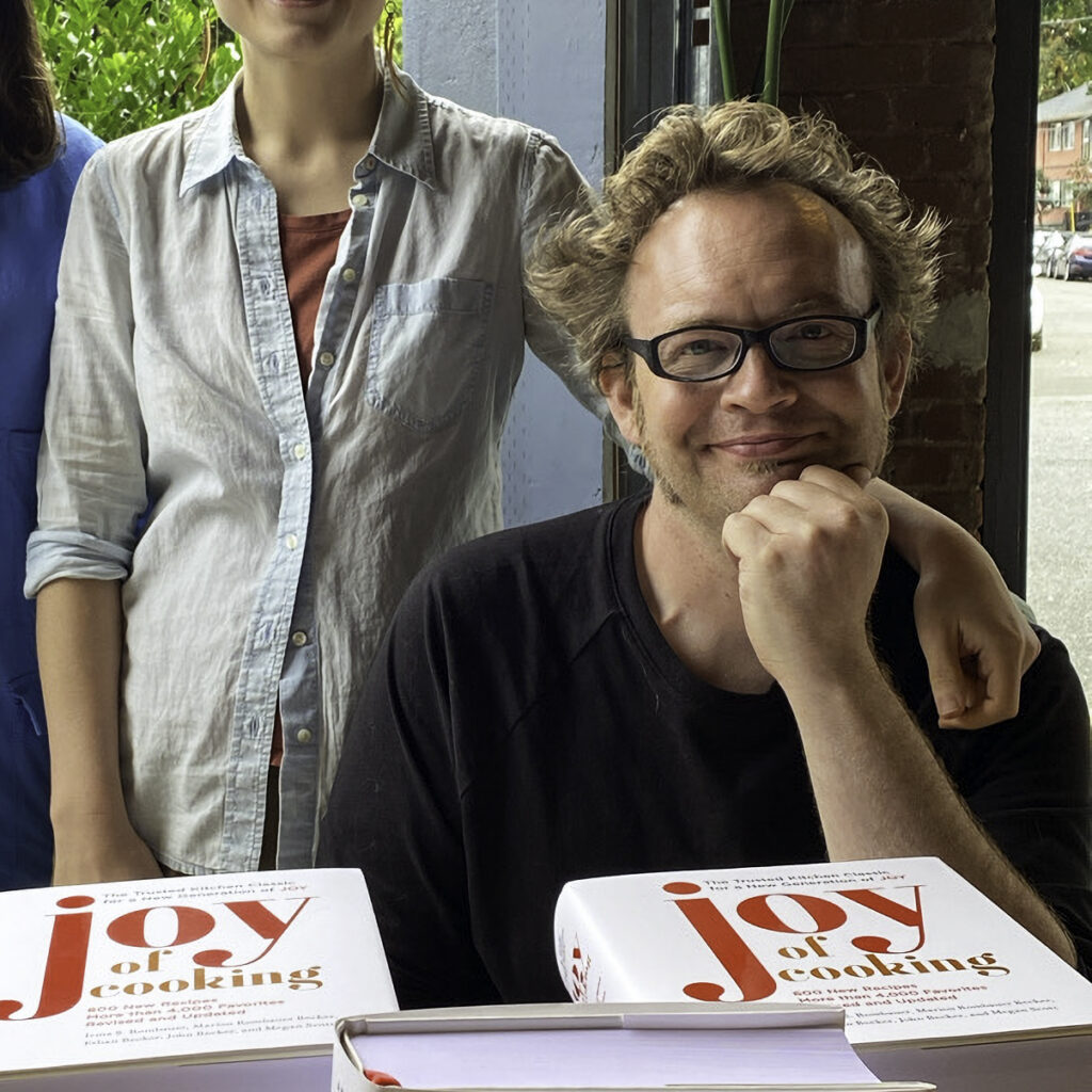 Portrait of John Becker, seated at a table behind several stacks of the 2019 edition of the Joy of Cooking. His head gently rests against an upturned arm. John is smiling genuinely, and not making a silly face, which is rare (and why this particular image has been chosen).