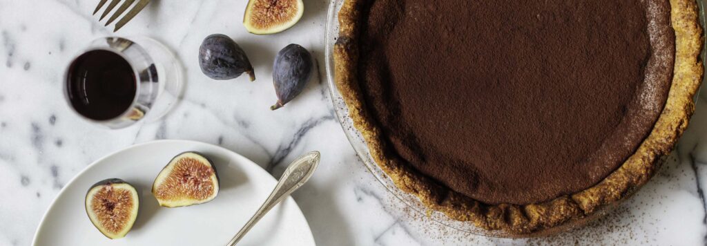 Overhead view of a chocolate chess pie served on a white marble surface with whipped cream and ripe, halved figs. A glass of port is nearby.