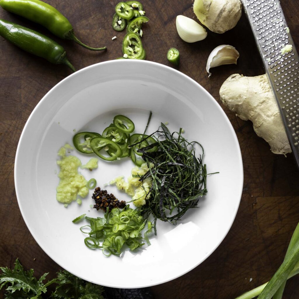Overhead view of a soup bowl containing grated ginger, grated garlic, thinly sliced serrano chile, thinly sliced mustard greens, and fried chile paste.