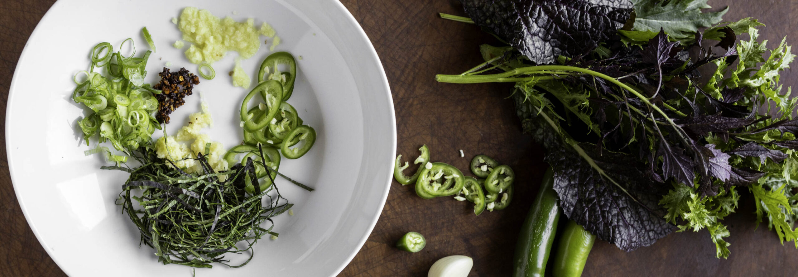 Overhead view of a soup bowl containing grated ginger, grated garlic, thinly sliced serrano chile, thinly sliced mustard greens, and fried chile paste. Mustard greens and whole serrano chiles are nearby.