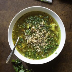 Overhead view of a bowl of chicken soup, topped with chopped herbs and fried shallots.