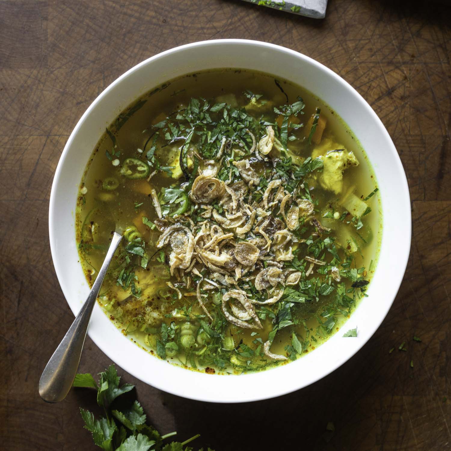 Overhead view of a bowl of chicken soup, topped with chopped herbs and fried shallots.