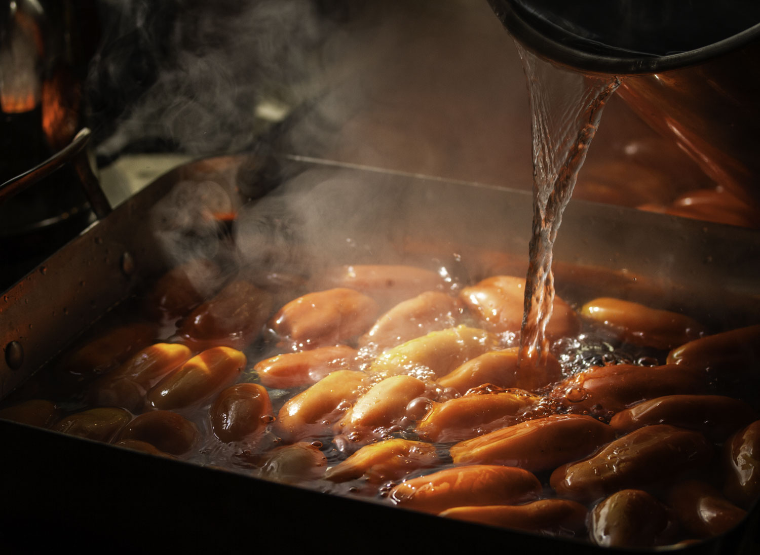 Moodily lit image of boiling water being poured into a deep roasting pan with full of roma tomatoes.