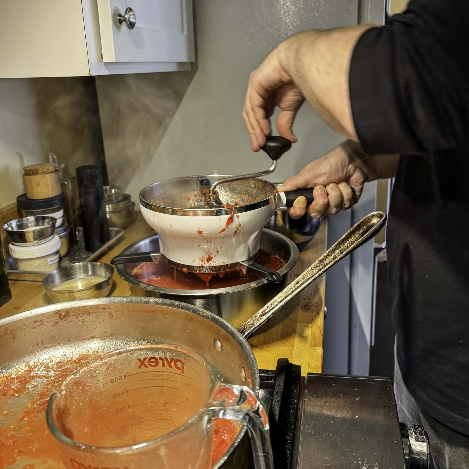 Simmered and mashed tomatoes are food milled by the author into a large stainless steel bowl. A nearly empty rondeau and a liquid measuring cup used for transferring the tomatoes is in the foreground.