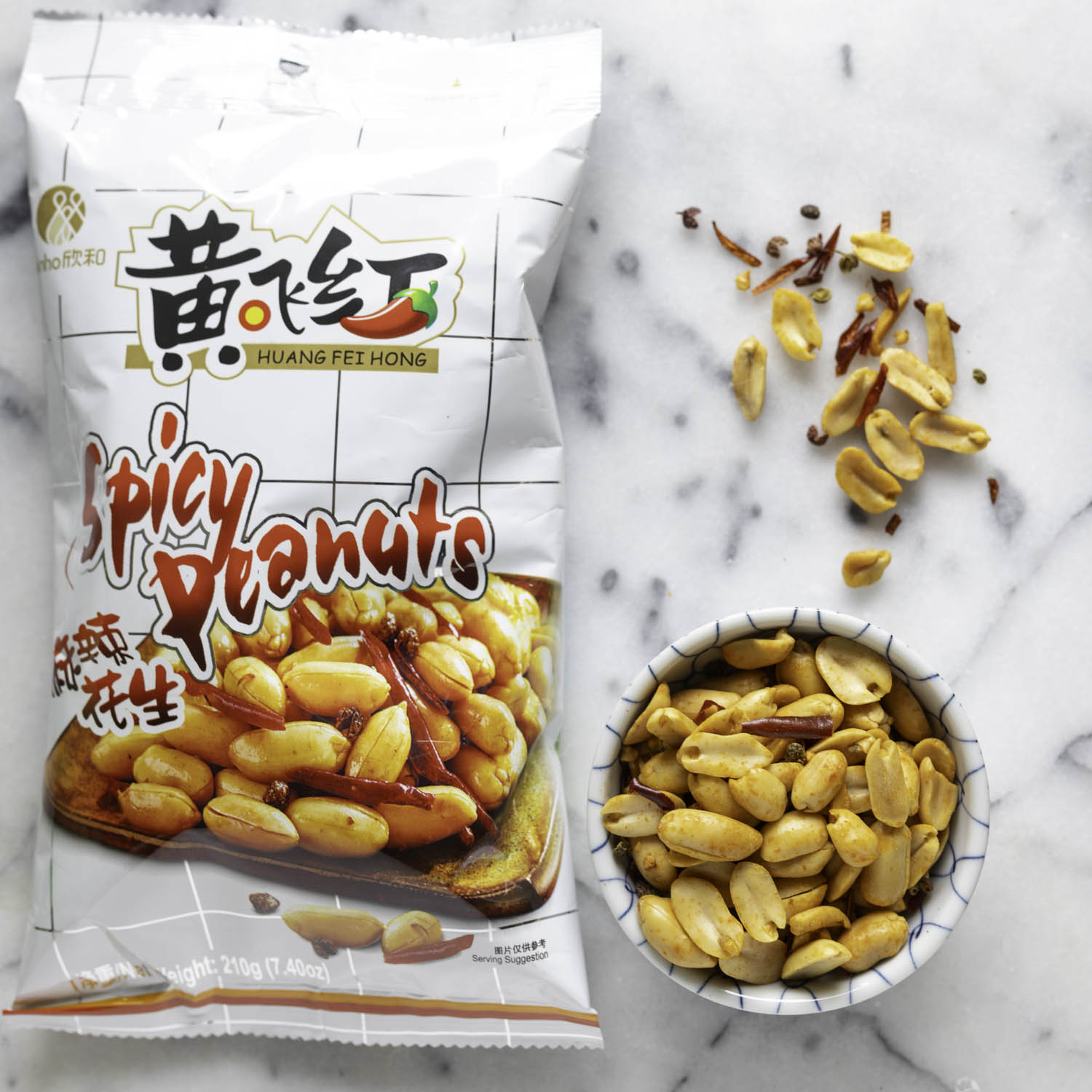 A bag of spicy peanuts, with a ramekin of the peanuts to one side. Some of the peanuts and spices have been strewn on the white marble surface.