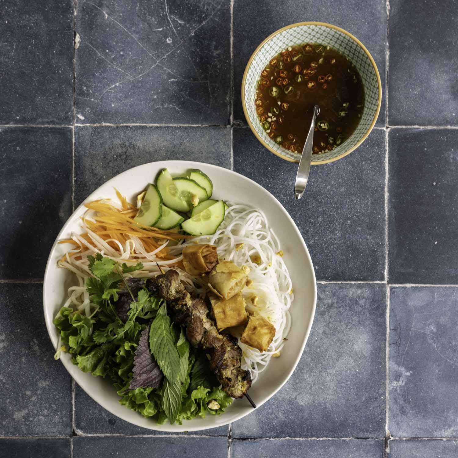 Overhead view of bún thit nuong, served with a small bowl of nuoc cham on a worn black tile surface.