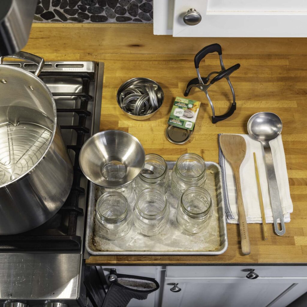 View of a stovetop and countertop. A stockpot filled with water is positioned over a burner near the counter edge. Jars, lids, towels, and various implements are arranged neatly on the counter.