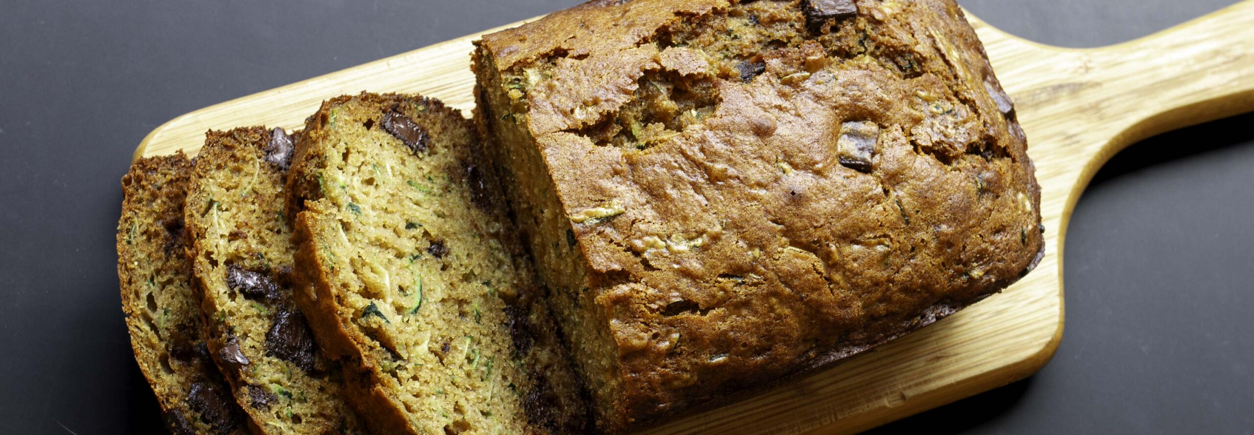 Overhead view of a loaf of chocolate zucchini bread. The has been placed on a small, handled cutting board. Half of the loaf has been cut into thick slices, which are shingled to reveal the tender, moist crumb.