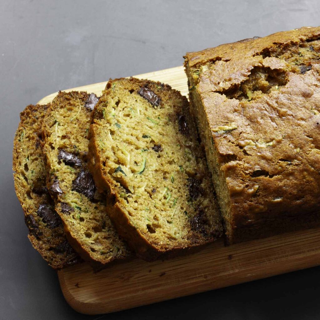 Overhead view of a loaf of chocolate zucchini bread. The has been placed on a small, handled cutting board. Half of the loaf has been cut into thick slices, which are shingled to reveal the tender, moist crumb.
