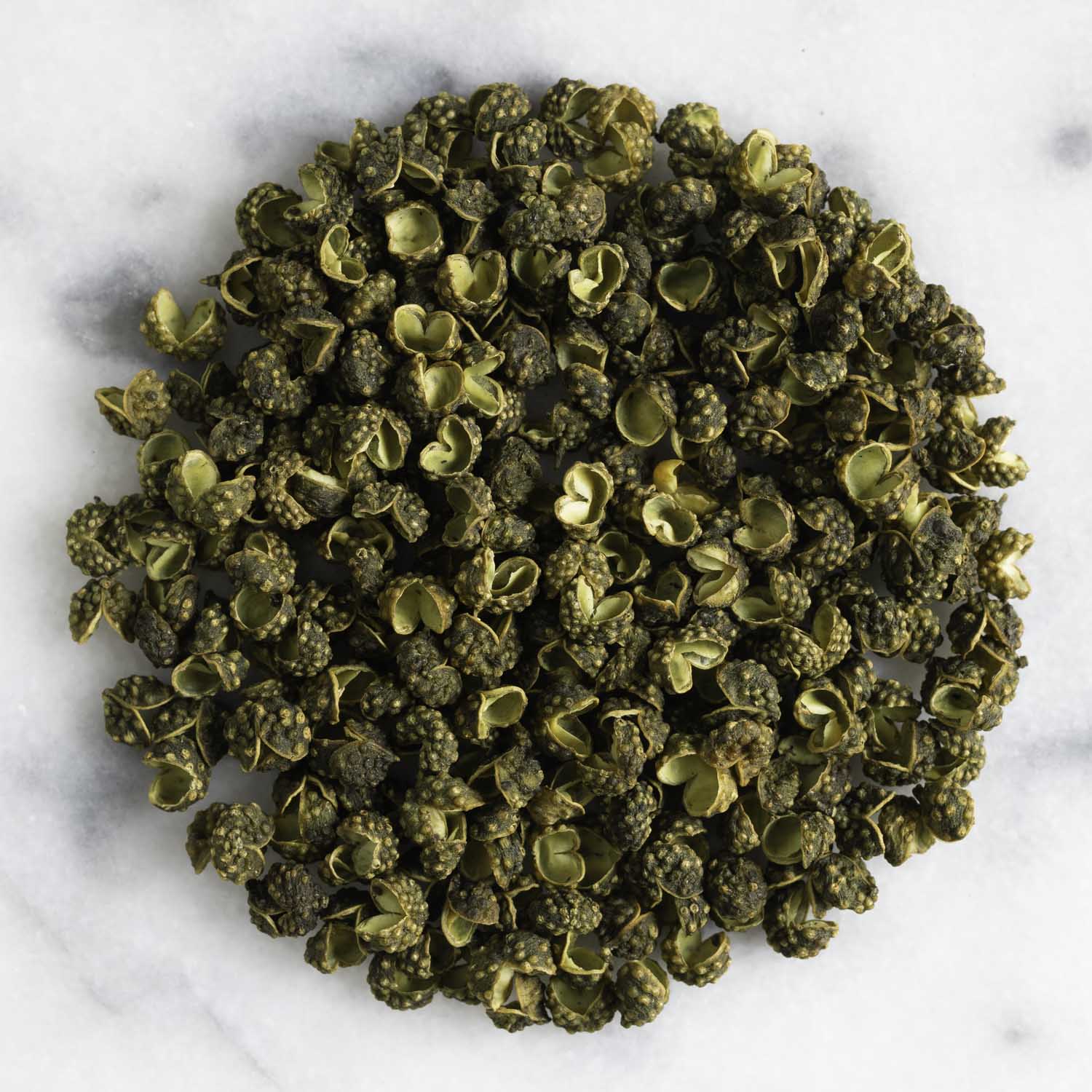 Closeup of a circular pile of dried green Sichuan pepper on a white marble surface.