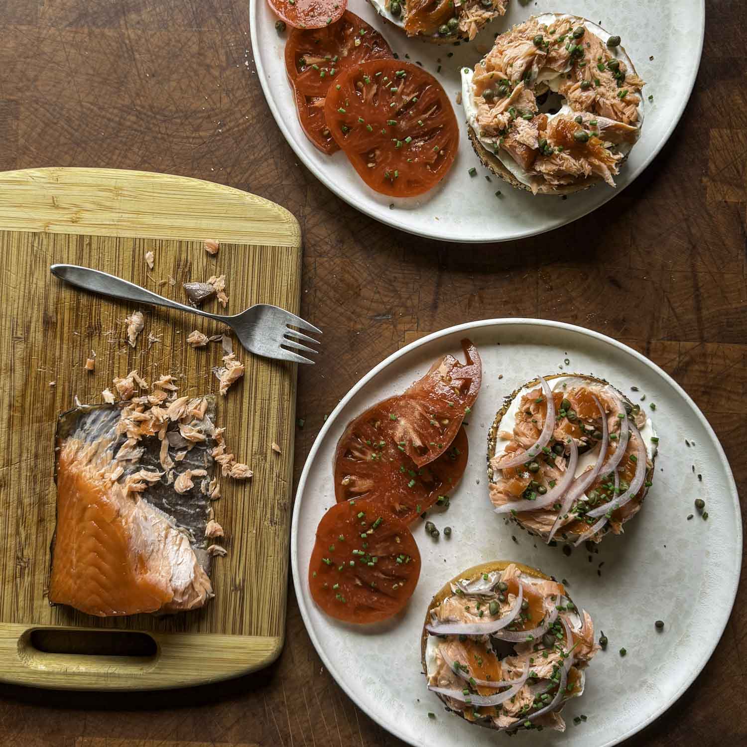 Two everything bagels served open face, topped with cream cheese, hot smoked salmon, sliced red onion, snipped chive, and capers. A cutting board of partially flaked hot-smoked salmon is nearby.