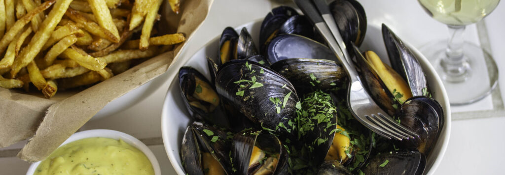 Steamed mussels, served with a bowl of fries and a ramekin of aîoli. A glass of white wine is nearby.