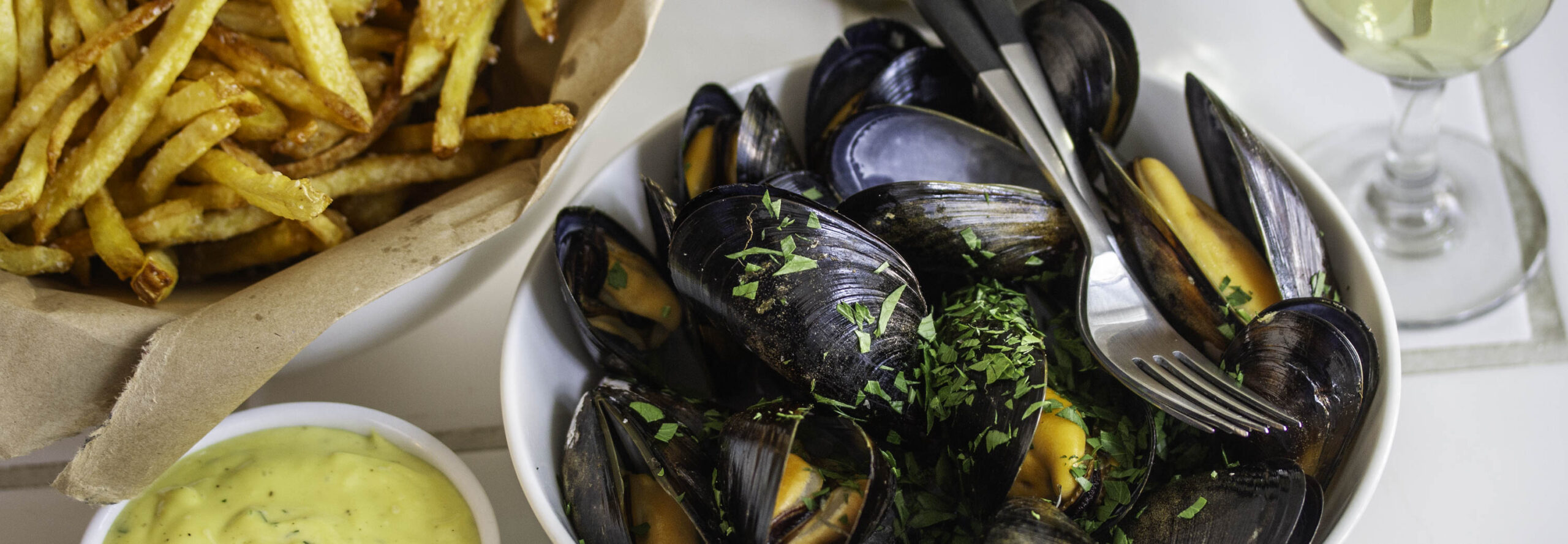 Steamed mussels, served with a bowl of fries and a ramekin of aîoli. A glass of white wine is nearby.