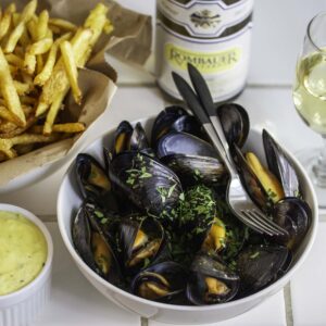 Steamed mussels, served with a bowl of fries and a ramekin of aîoli. A glass of white wine is nearby.