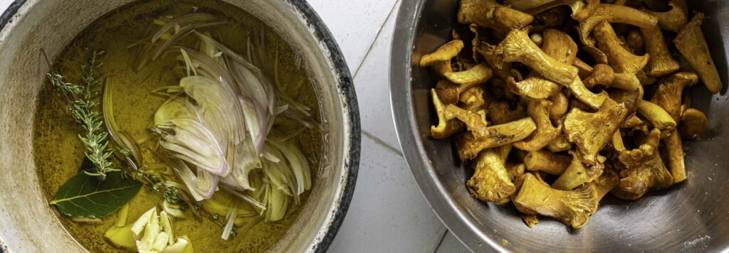 Overhead view of salted chanterelle mushrooms in a mixing bowl. A Dutch oven containing olive oil and garlic, shallot, and fresh herbs is nearby.