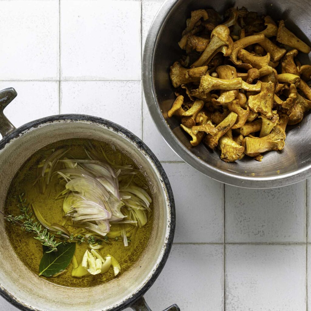 Overhead view of salted chanterelle mushrooms in a mixing bowl. A Dutch oven containing olive oil and garlic, shallot, and fresh herbs is nearby.