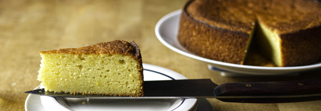 Profile view of a slice of olive oil cake perched on the blade of a chef's knife over a small serving plate. The rest of the cake is visible in the background, slightly out of focus.