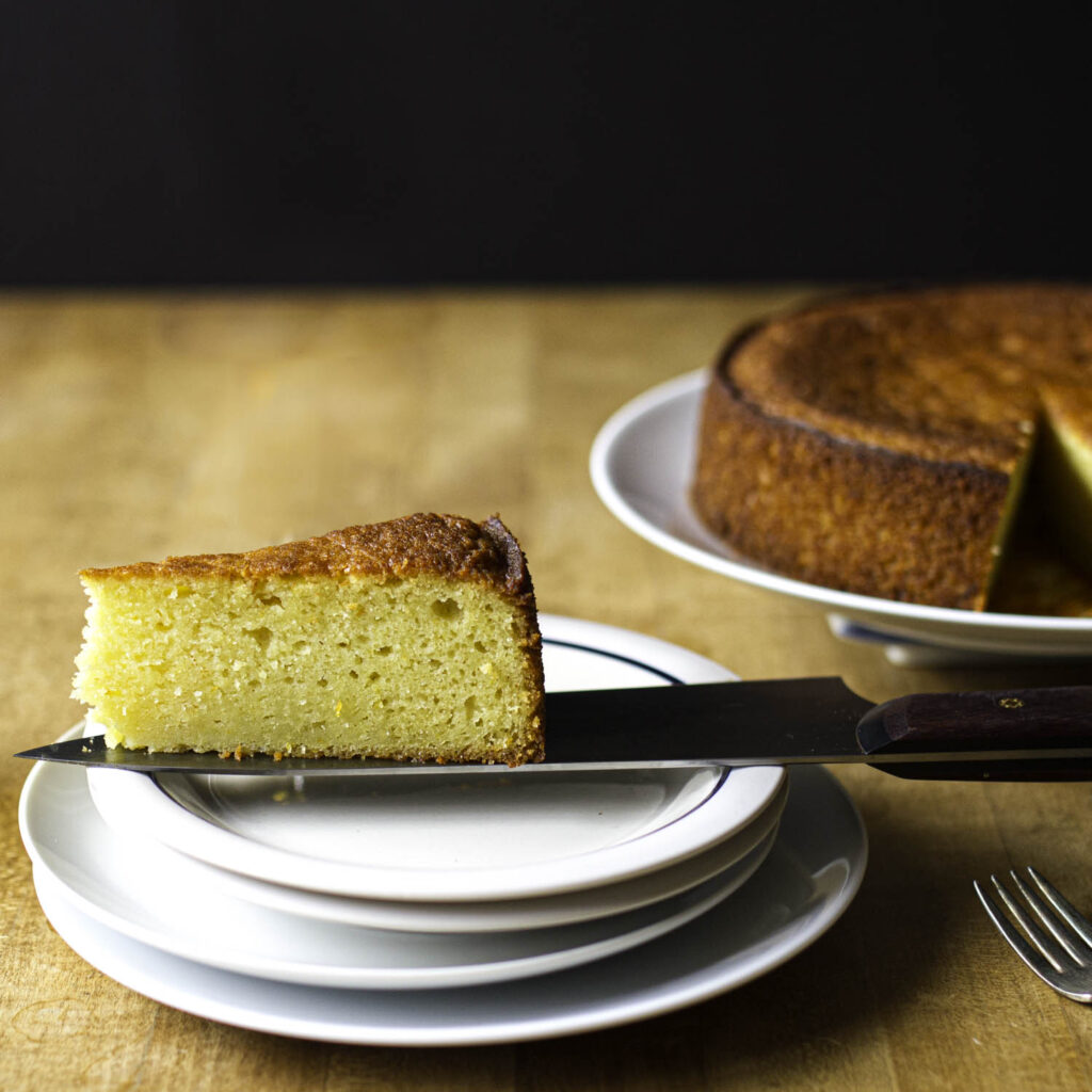Profile view of a slice of olive oil cake perched on the blade of a chef's knife over a small serving plate. The rest of the cake is visible in the background, slightly out of focus.