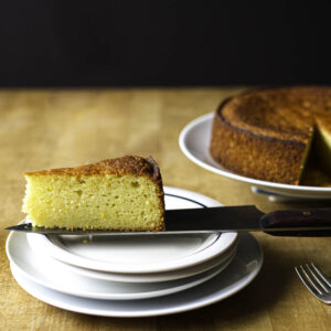 Profile view of a slice of olive oil cake perched on the blade of a chef's knife over a small serving plate. The rest of the cake is visible in the background, slightly out of focus.
