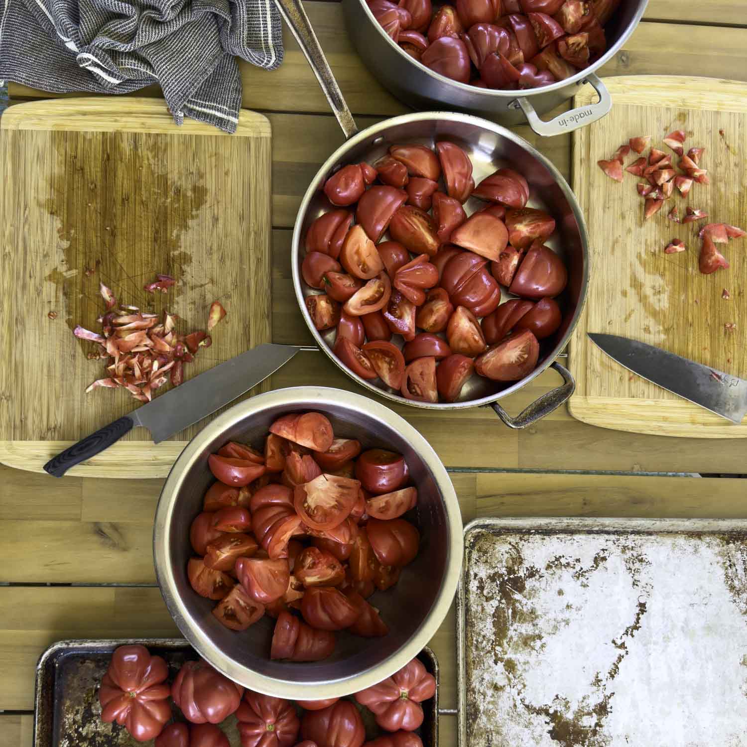 Overhead view of two prep stations with cutting boards and chef's knives. Two pans and a bowl are full of quartered and cored tomatoes.