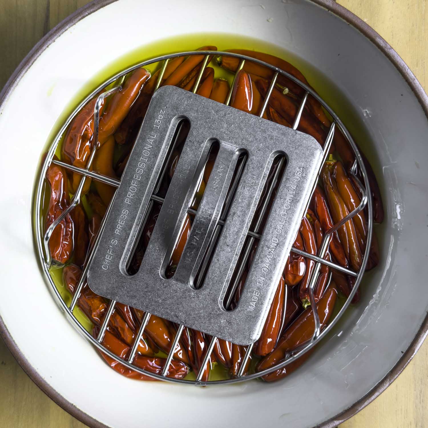 The salted, pickled, and dried chiles are submerged in olive oil in a bowl. A round wire rack and cooking press have been placed on top to keep them covered with the oil.