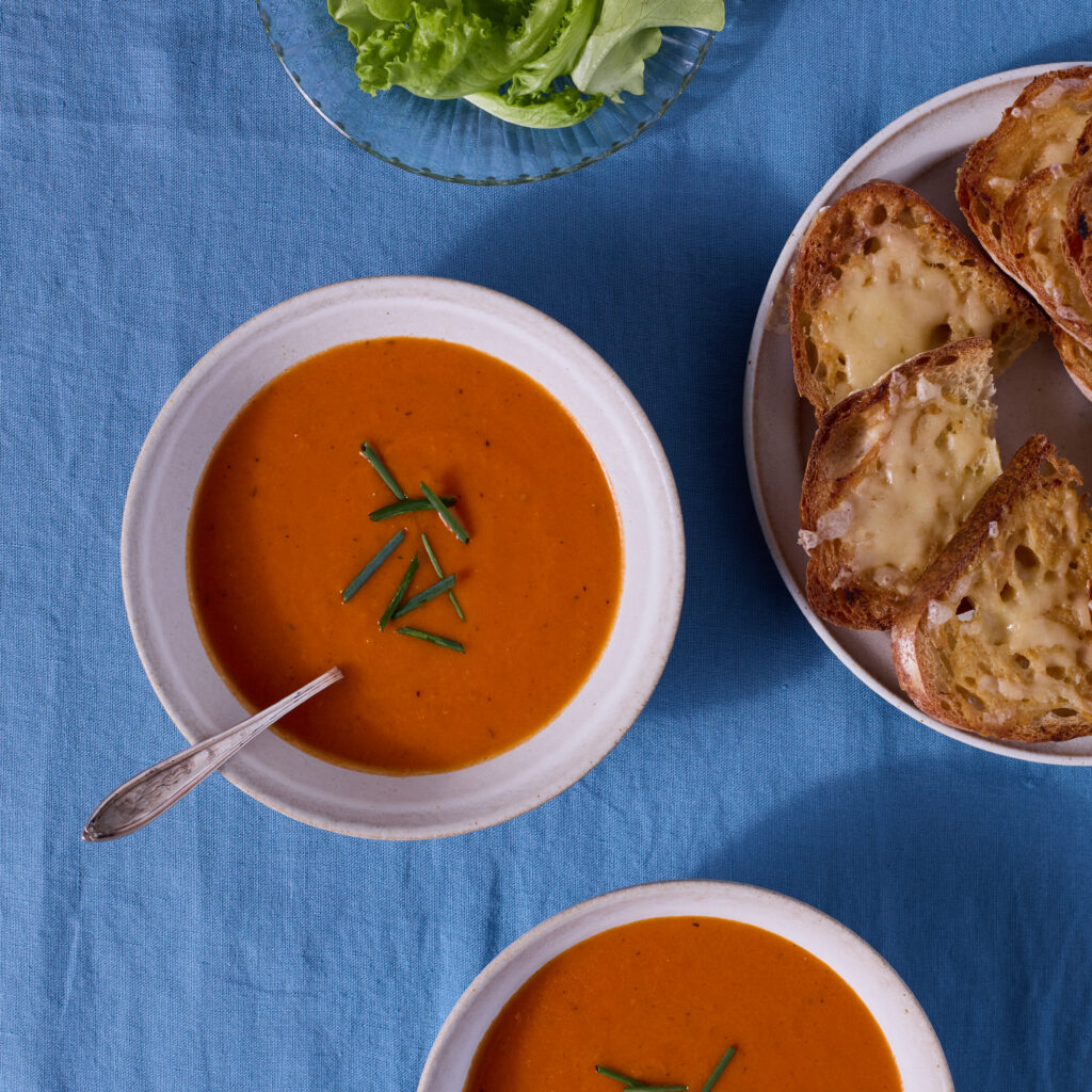Two bowls of tomato-orange soup are served on a blue fabric surface. A plate of cheese toasts and a bowl of dressed salad greens are nearby.