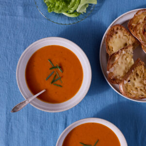 Two bowls of tomato-orange soup are served on a blue fabric surface. A plate of cheese toasts and a bowl of dressed salad greens are nearby.