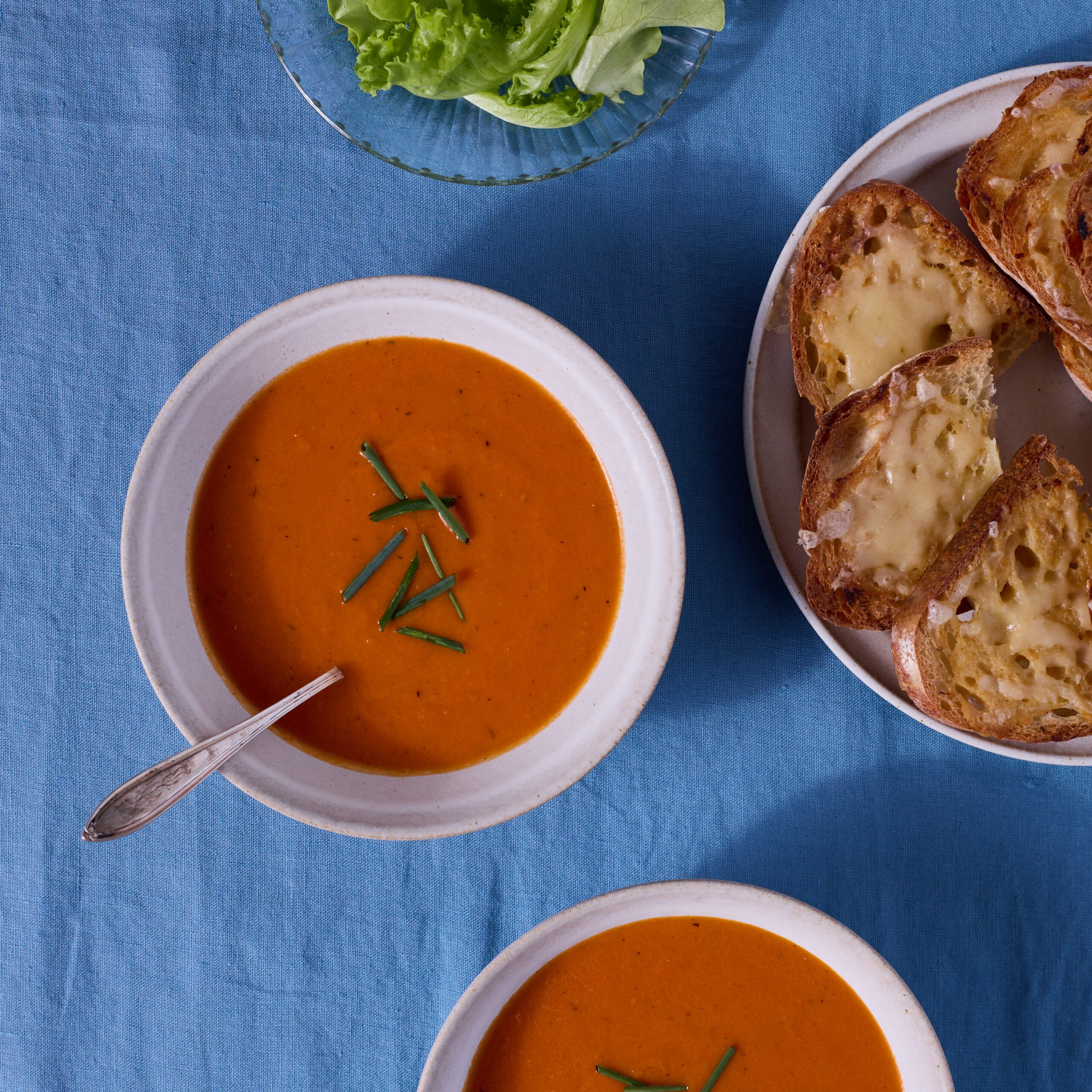 Two bowls of tomato-orange soup are served on a blue fabric surface. A plate of cheese toasts and a bowl of dressed salad greens are nearby.