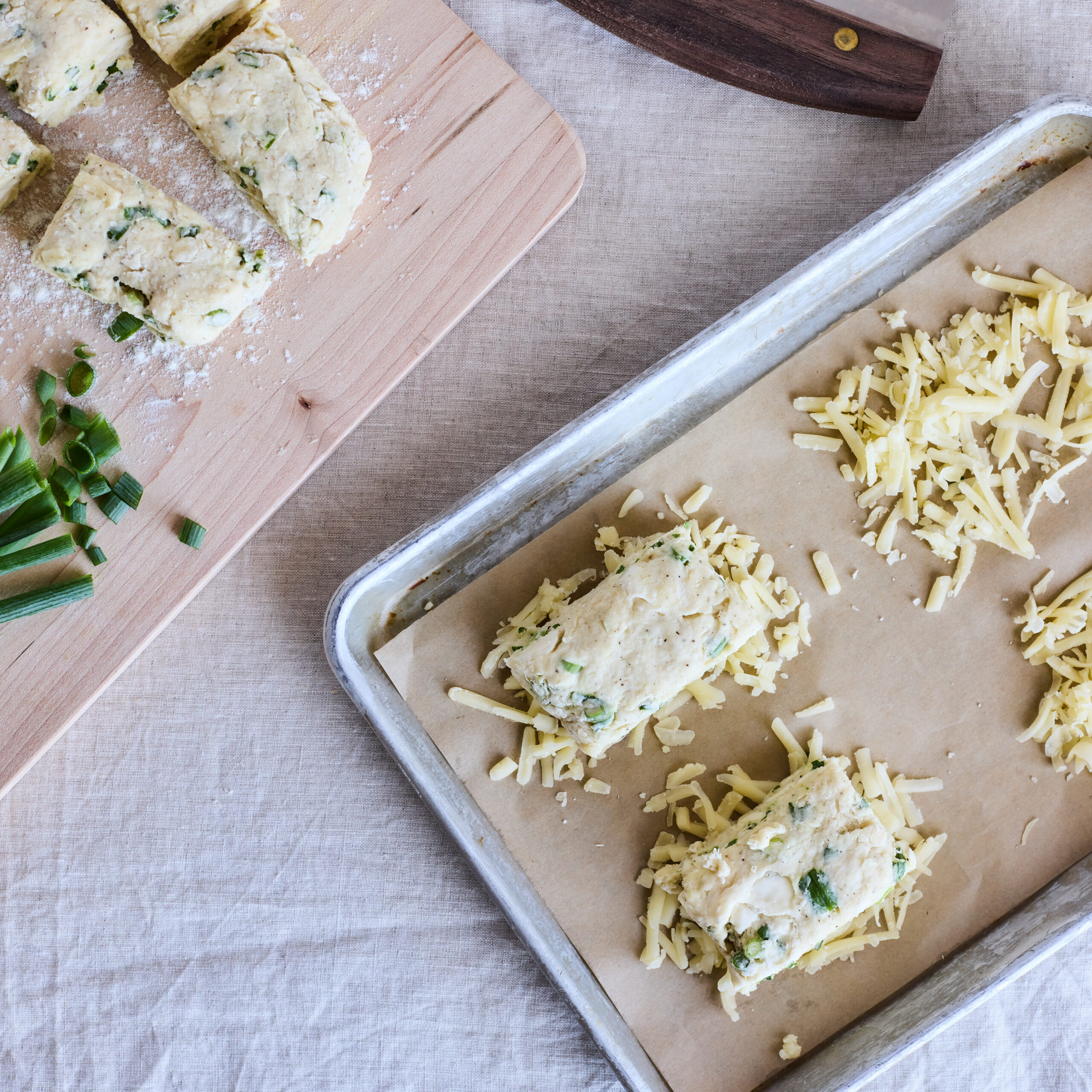 Overhead view of a parchment lined sheetpan with four thin mounds of shredded cheese. Two cut portions of cheddar-scallion biscuit dough have been placed on two of these cheese mounds.