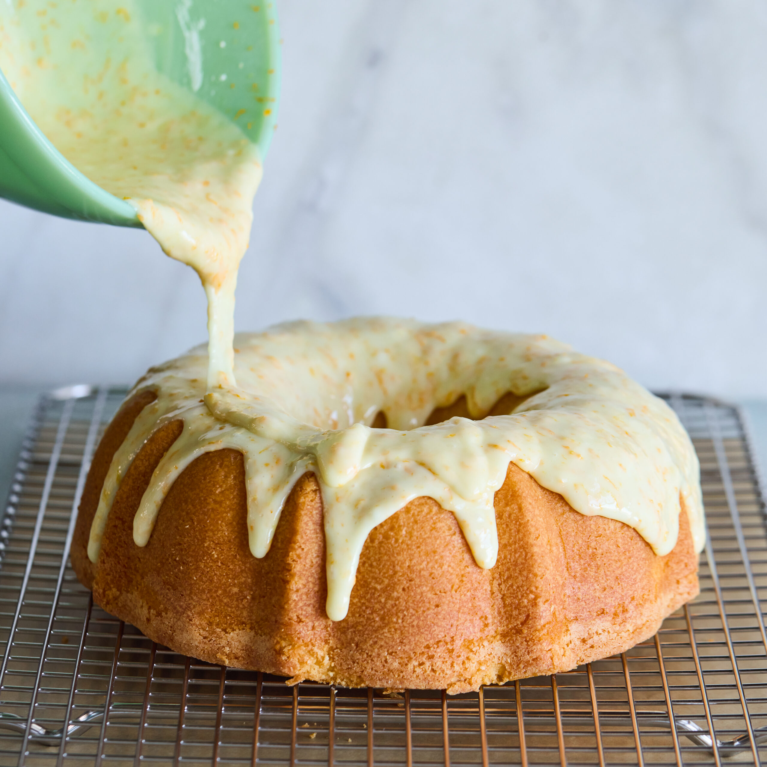 A mimosa pound cake set on a cooling rack. A thick orange-sparkling wine glaze is being poured over the pound cake from a jadeite batter bowl.